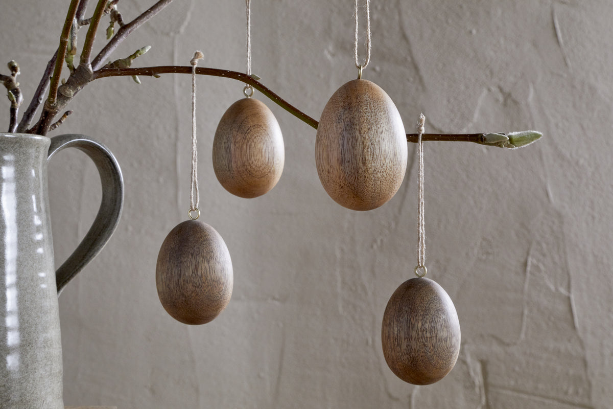 Nkuku’s Dima Mango Wood Hanging Egg Decorations (Set of 4) add minimalist, natural Easter décor when hung from a branch in a gray ceramic pitcher, set against a textured neutral-toned wall.