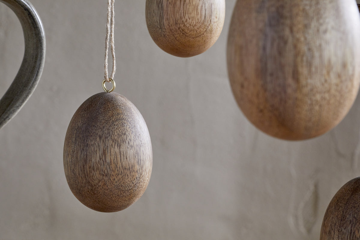 The nkuku Dima Mango Wood Hanging Egg Decoration – Natural (Set of 4) features smooth, brown mango wood eggs suspended on strings for a natural, minimalist Easter accent against textured beige walls.
