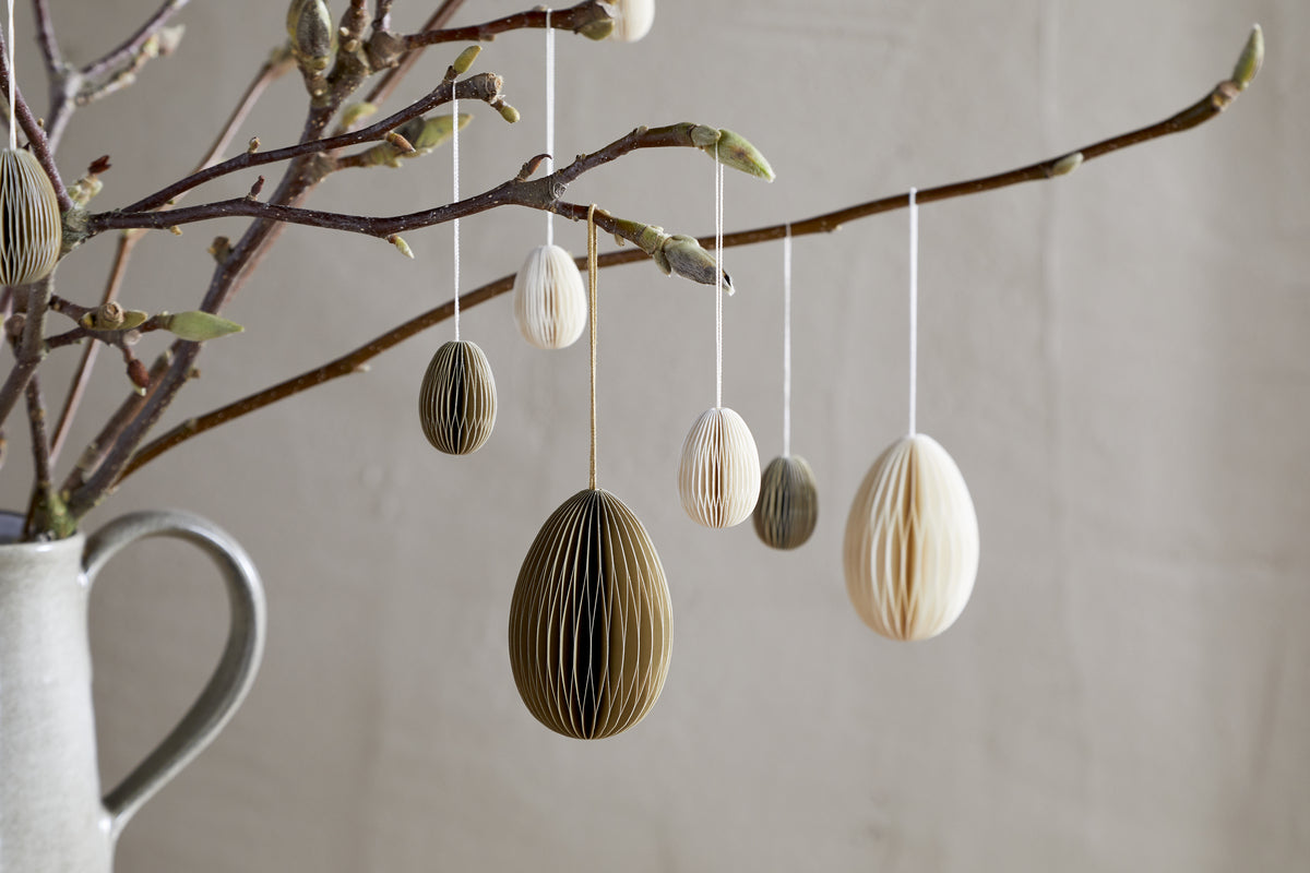 A ceramic jug holds bare branches from which nkuku’s moss green Piani Hanging Paper Decorations (set of 4) are suspended, creating a minimalist and natural Easter display against a plain beige background.