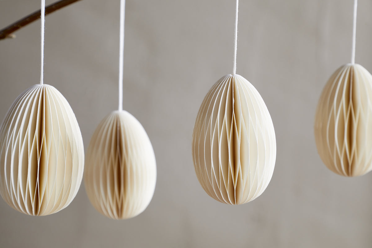 The nkuku Piani Hanging Paper Decoration - Cream - Set of 4 features cream-colored honeycomb eggs suspended vertically on white strings, each displaying a delicate geometric pleated texture and soft shadows.