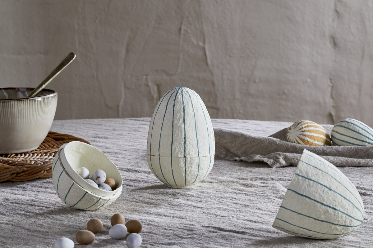 A textured Easter table features nkuku’s Surya Recycled Cotton Paper Standing Opening Egg in teal & cream, with one open to reveal candy eggs inside. Two smaller striped eggs and a bowl with a spoon sit in the background on the left.