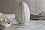 The Surya Recycled Cotton Paper Standing Opening Egg in teal and cream by nkuku sits on a beige cotton paper surface. In front, a partial bowl holds speckled eggs, with neutral cloth and a blurred round object in the background.