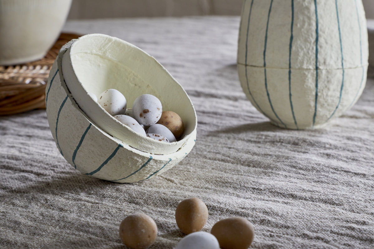 The nkuku Surya Recycled Cotton Paper Standing Opening Egg in teal & cream is displayed on a beige linen surface, spilling small speckled eggs from its opening. A matching container and wicker tray sit in the background, perfect for Easter décor.