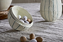 The nkuku Surya Recycled Cotton Paper Standing Opening Egg in teal & cream is displayed on a beige linen surface, spilling small speckled eggs from its opening. A matching container and wicker tray sit in the background, perfect for Easter décor.