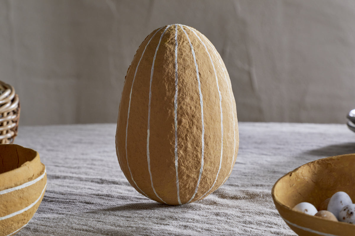 The nkuku Surya Recycled Cotton Paper Side Opening Egg in ochre & cream stands upright on a light cloth, making a striking Easter table centerpiece. Two yellow bowls with white eggs and a woven basket complete the seasonal display.