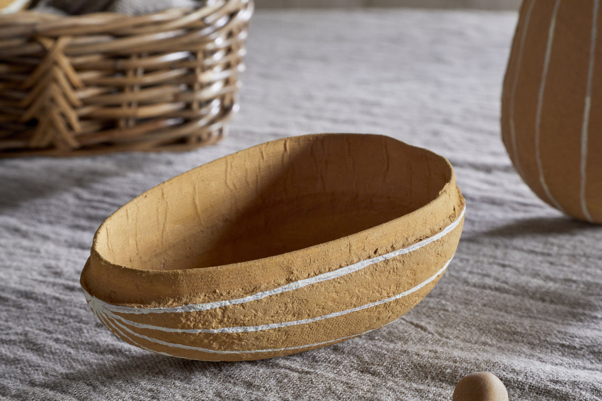 The Surya Recycled Cotton Paper Side Opening Egg in ochre & cream by nkuku rests on beige fabric, its handmade design with white stripes ideal for Easter tables. Behind it, a wicker basket and another striped ceramic piece are visible.