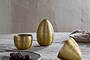 Three Mutti Hammered Opening Eggs in antique brass by nkuku, with intricate engraving, sit on a textured light gray tablecloth. One is open to reveal its interior. A plate of dark treats and a beige bowl with gray napkin are nearby.