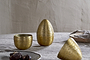 Three Mutti Hammered Opening Eggs in antique brass by nkuku, with intricate engraving, sit on a textured light gray tablecloth. One is open to reveal its interior. A plate of dark treats and a beige bowl with gray napkin are nearby.