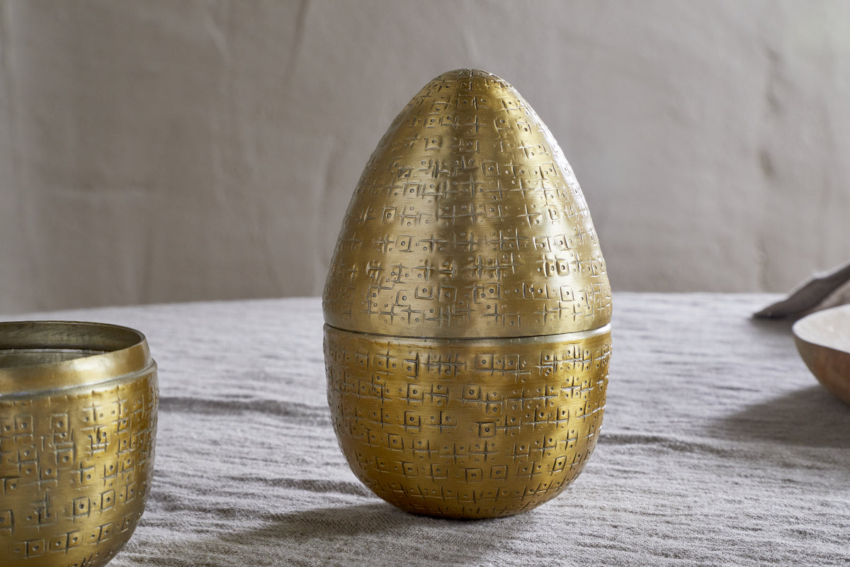 The Mutti Hammered Opening Egg in antique brass by nkuku, with intricate geometric engravings, rests on a light gray textured cloth. A matching round bowl sits to the left and a blurred tan dish appears in the right background.