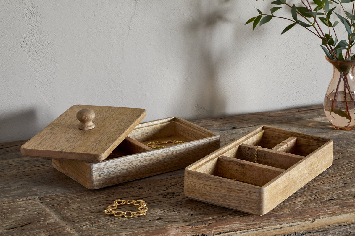 Two nkuku Chanya Mango Wood Jewellery Boxes in natural finish sit on a rustic table; one hand-carved box is open, displaying gold jewelry, while a gold bracelet and a glass vase with greenery complete the scene.