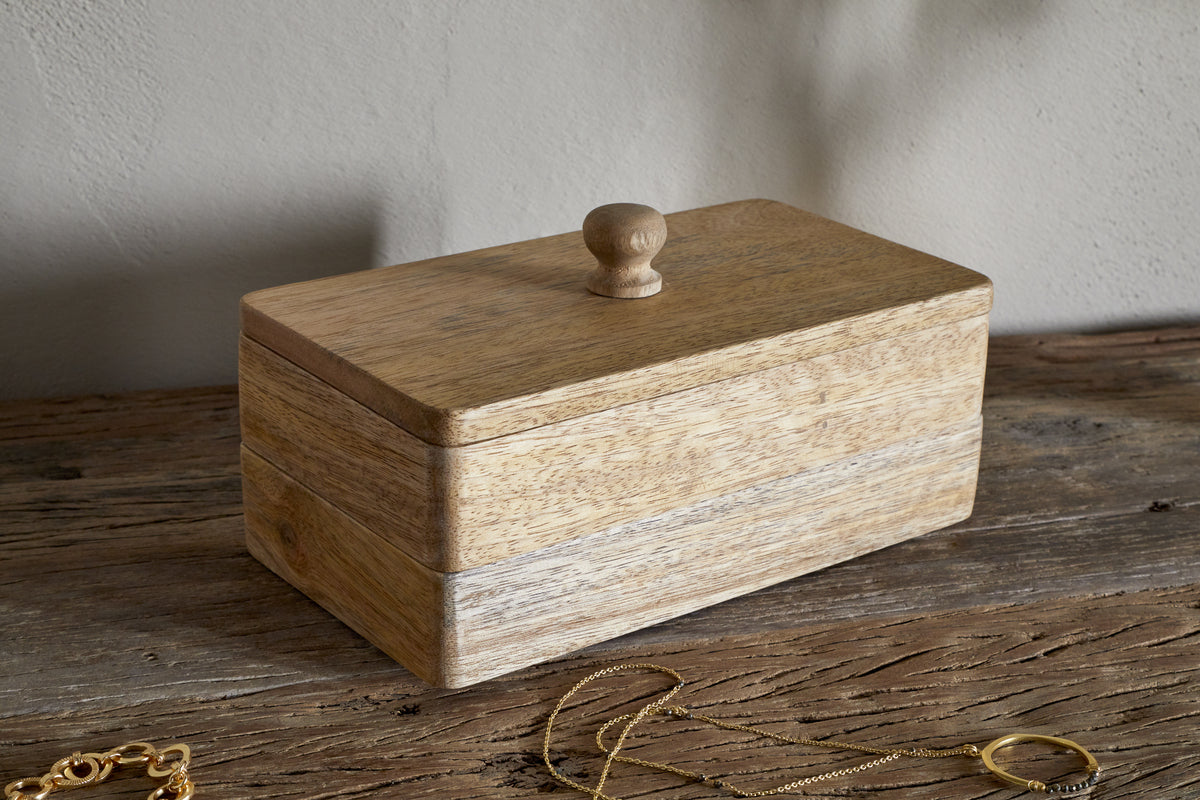 The Chanya Mango Wood Jewellery Box - Natural by nkuku, a rectangular hand-carved piece with a round lid knob, rests on a rustic surface near gold jewelry. The background features a plain, light-colored wall.