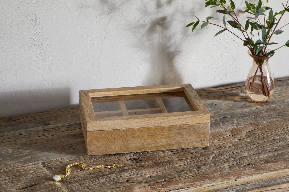 The Kabani Mango Wood Jewellery Box - Natural by nkuku rests on a rustic table, its glass lid revealing empty compartments. Beside it lies a gold bracelet with a charm, and a vase of green stems completes the scene.