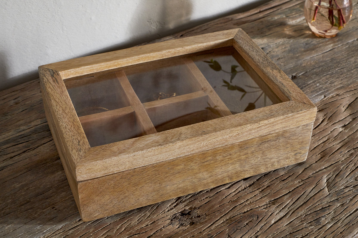 The nkuku Kabani Mango Wood Jewellery Box - Natural, featuring a glass lid and four compartments, showcases its visible wood grain as it rests on a rustic table. Light reflects on the glass, with part of a vase in the top right corner.