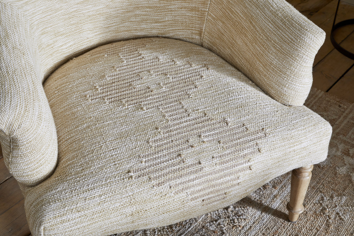 Close-up of the nkuku Aranya Cotton & Jute Occasional Chair in Natural, featuring a geometric pattern, rounded arms, and wooden legs. The chair sits on a patterned light brown rug atop a wooden floor.
