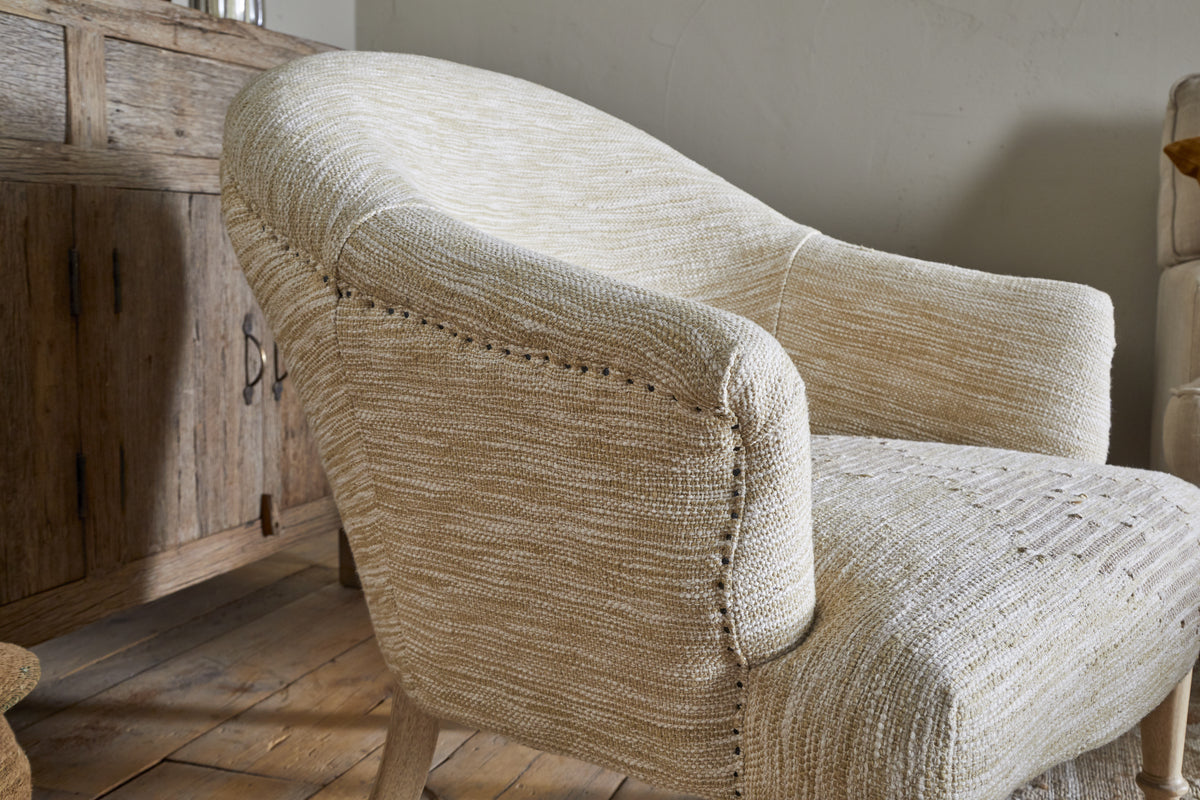 A close-up of the Aranya Cotton & Jute Occasional Chair - Natural by nkuku shows its textured beige fabric, rounded arms and back with nailhead trim. The chair sits on light wood flooring beside a rustic cabinet, creating a cozy setting.