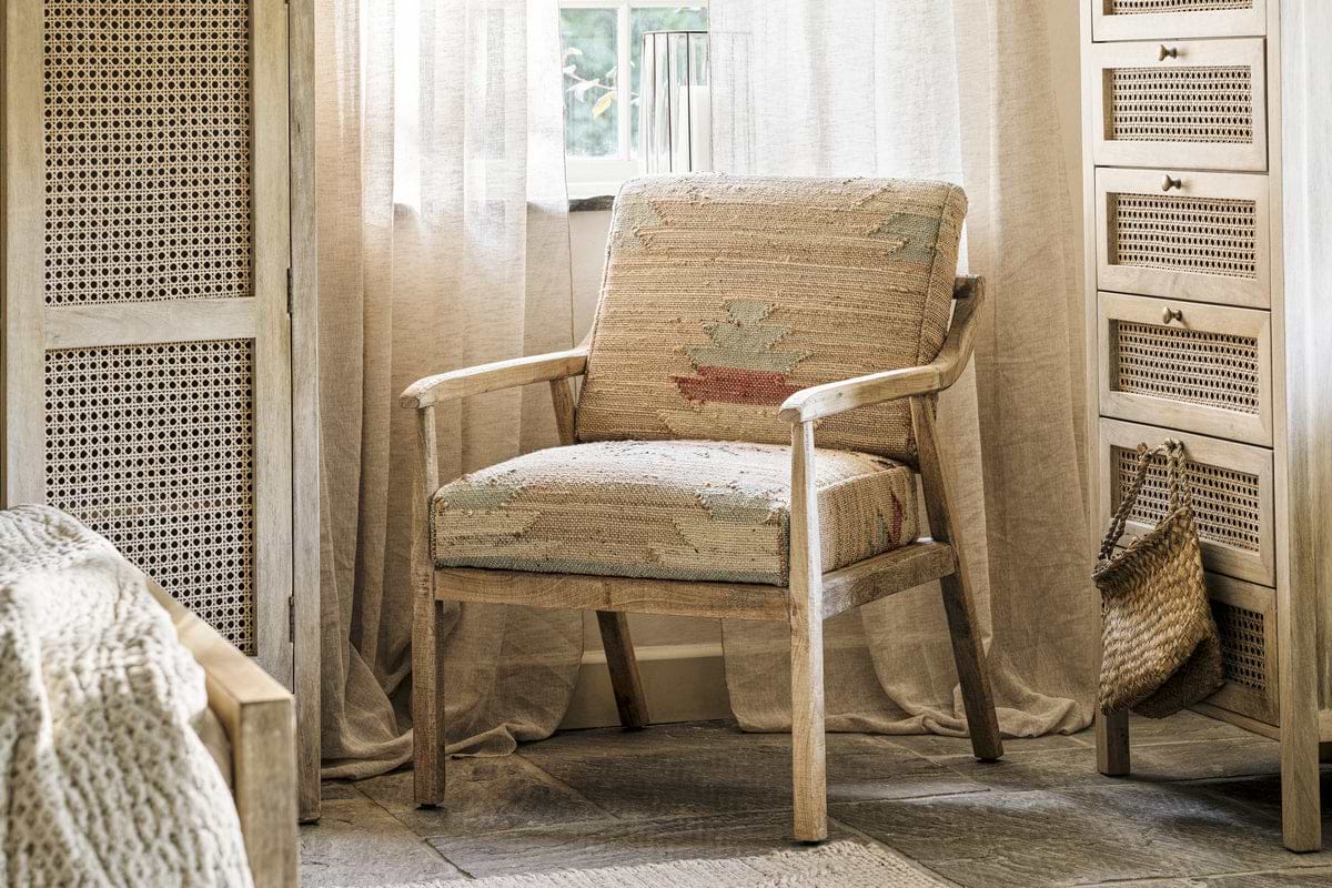 The Kavya Kilim Occasional Armchair by nkuku features natural, patterned upholstery and sits in a cozy room with woven accents, sheer curtains, a wicker bag on the cabinet, and sunlight streaming in for a warm ambiance.
