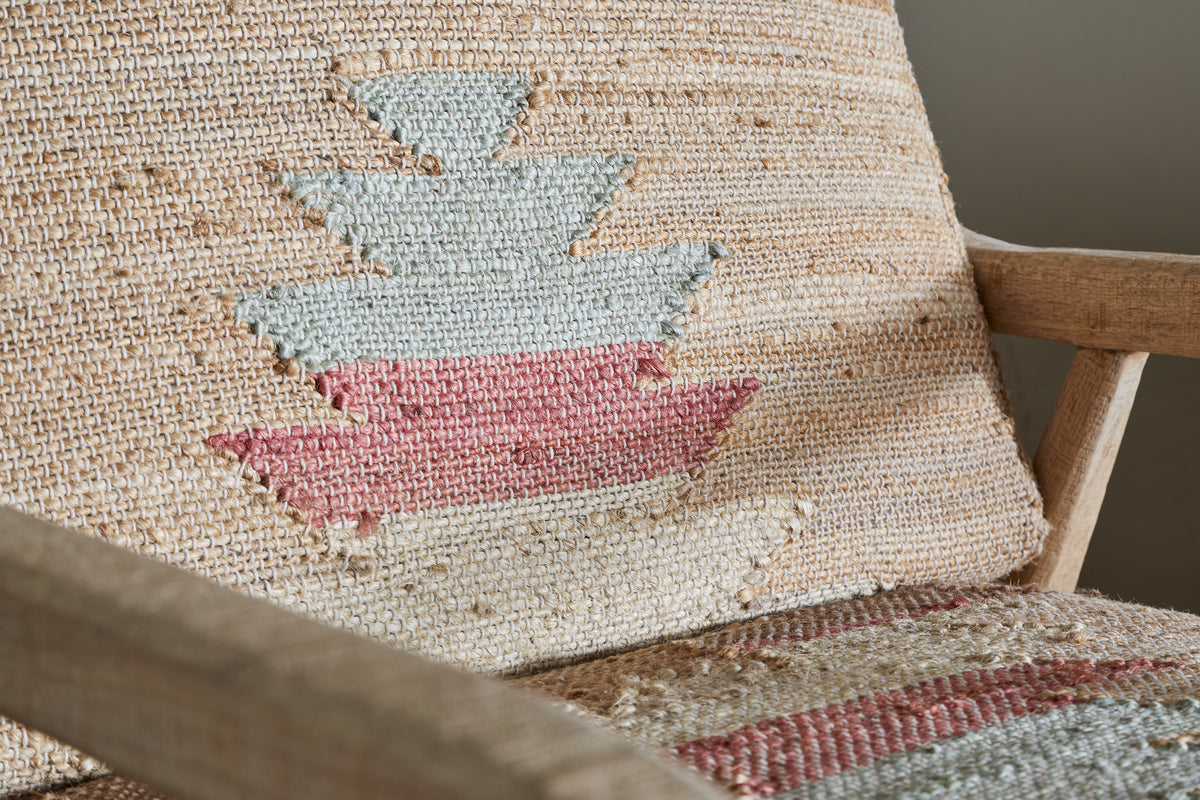 Close-up of the Kavya Kilim Occasional Armchair by nkuku, featuring a woven fabric with a geometric pattern in beige, light blue, and pink, plus visible wooden armrest and backrest with a textured natural finish.