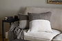 A beige sofa with two square pillows, one in the nkuku Munir Linen Cushion Cover—charcoal with gray and off-white color blocks. A gray knit throw rests on the armrest; nearby, a dark side table holds stacked books and a mug against a light wall.