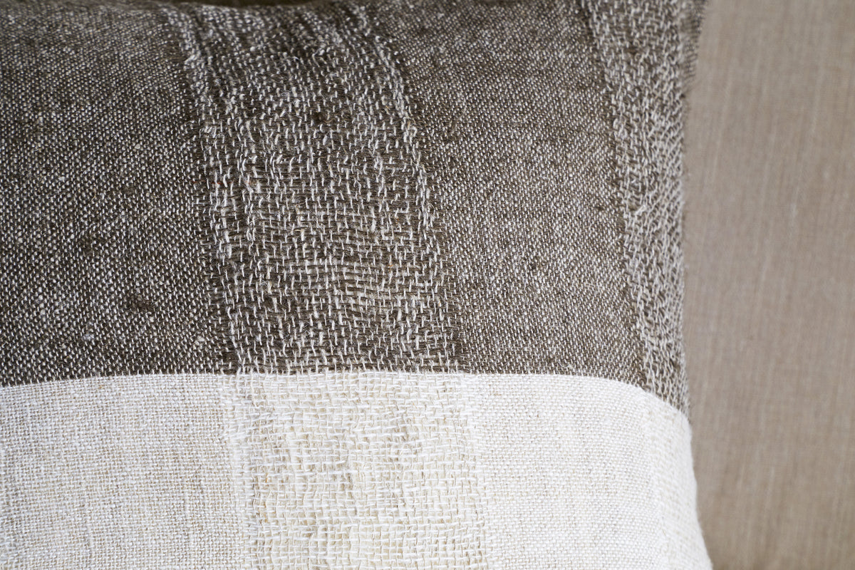 A close-up of the nkuku Munir Linen Cushion Cover - Charcoal shows its slubby textured linen and horizontal color block design—brownish-gray on top, off-white below—set against a beige upholstered background.