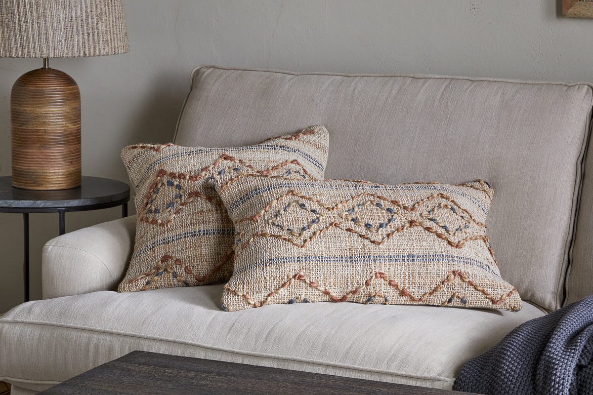 A beige sofa features two nkuku Nila Esha Cushion Covers in rust and indigo geometric patterns. A round black side table holds a brown ribbed lamp with a beige shade, while a blue knitted blanket is draped on the sofa’s armrest.