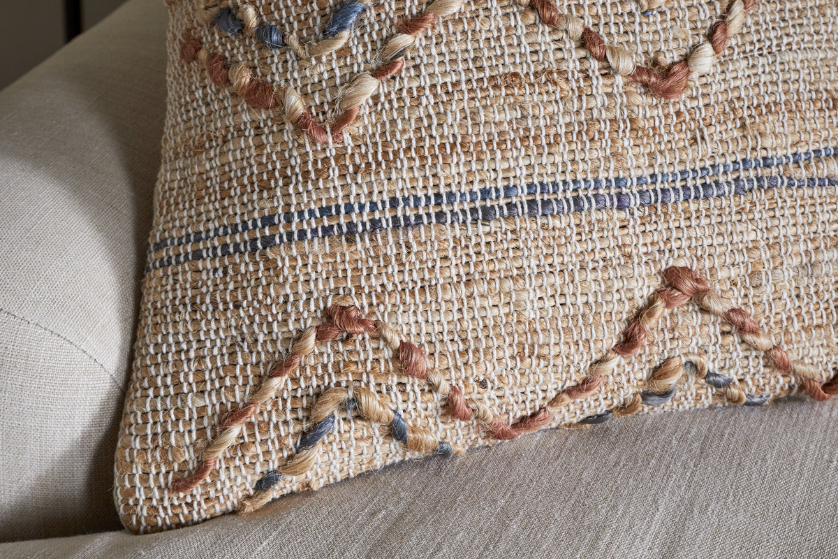 The Nila Esha Cushion Cover by nkuku features textured zigzag and horizontal patterns in rust and indigo, with a natural, handmade look. Shown here on a beige upholstered chair, its woven fabric adds rustic charm to any space.