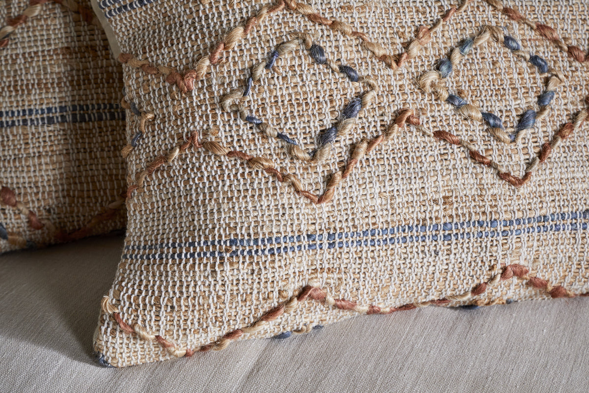Close-up of the hand-embroidered nkuku Nila Esha Cushion Cover – Rust & Indigo, featuring textured diamond and line patterns on a beige base. It sits on a light beige surface with another similar cushion nearby.