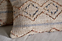 Close-up of the hand-embroidered nkuku Nila Esha Cushion Cover – Rust & Indigo, featuring textured diamond and line patterns on a beige base. It sits on a light beige surface with another similar cushion nearby.
