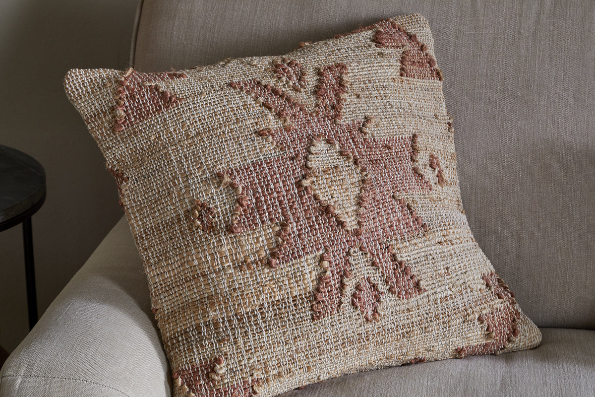 The nkuku Nila Gul Cushion Cover - Rust features a geometric, tribal-inspired pattern in earthy beige and muted rust tones, crafted from cotton and jute for a textured, rustic look that complements a light beige sofa.