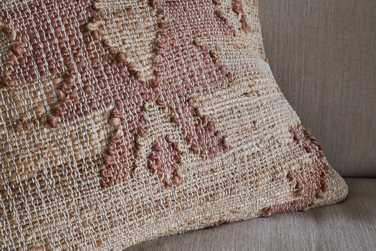 The Nila Gul Cushion Cover - Rust by nkuku showcases a geometric, southwestern-inspired pattern in earthy beige and muted rust tones. Its textured cotton and jute weave creates a rustic, artisanal look, perfect for accenting a taupe fabric couch.