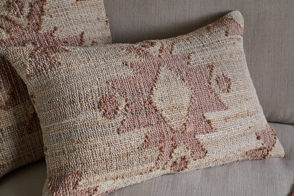 The nkuku Nila Gul Cushion Cover in Rust, featuring a textured geometric pattern on woven beige cotton and jute, adds a handmade, rustic touch. One rests on a matching beige couch with another similar cushion partially visible behind it.