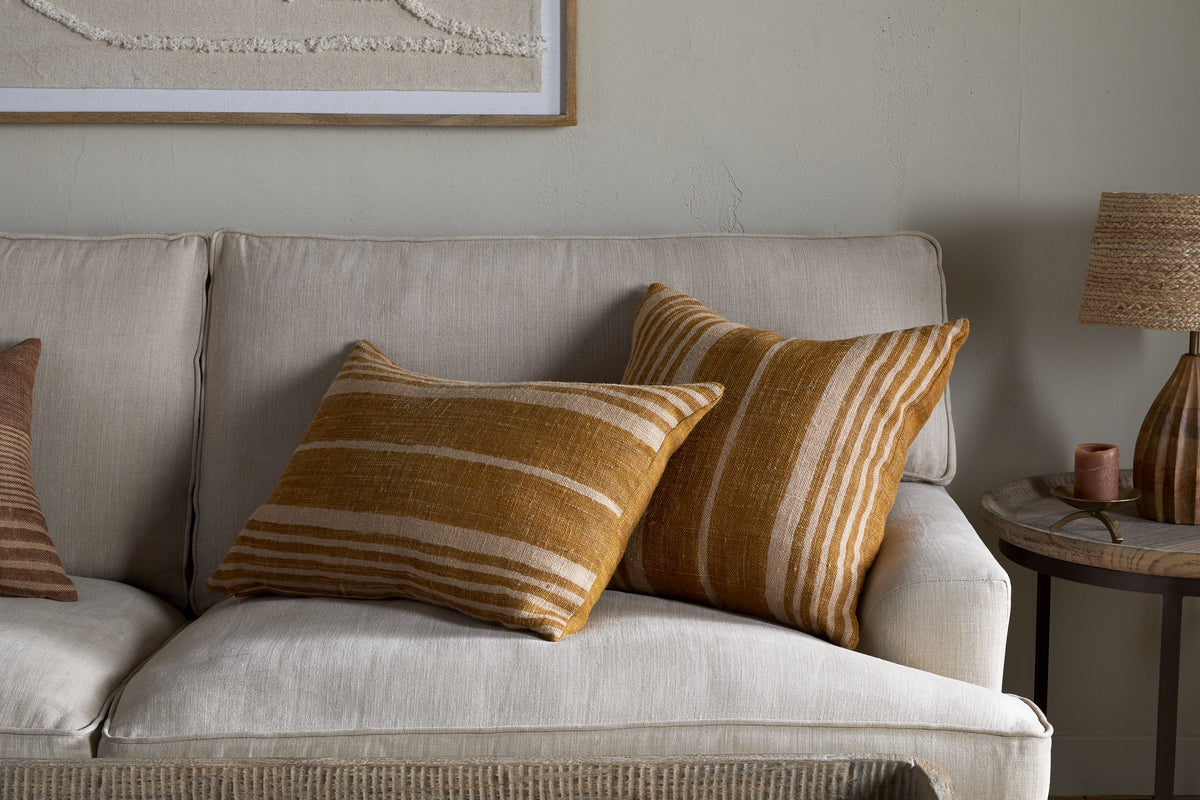 A beige sofa is styled with nkuku's Eesha Jute Cushion Cover in ochre and striped pillows for a cozy look. A brown and cream block-printed cushion cover sits to the left. A wooden side table holds a woven lamp and decor under framed artwork.