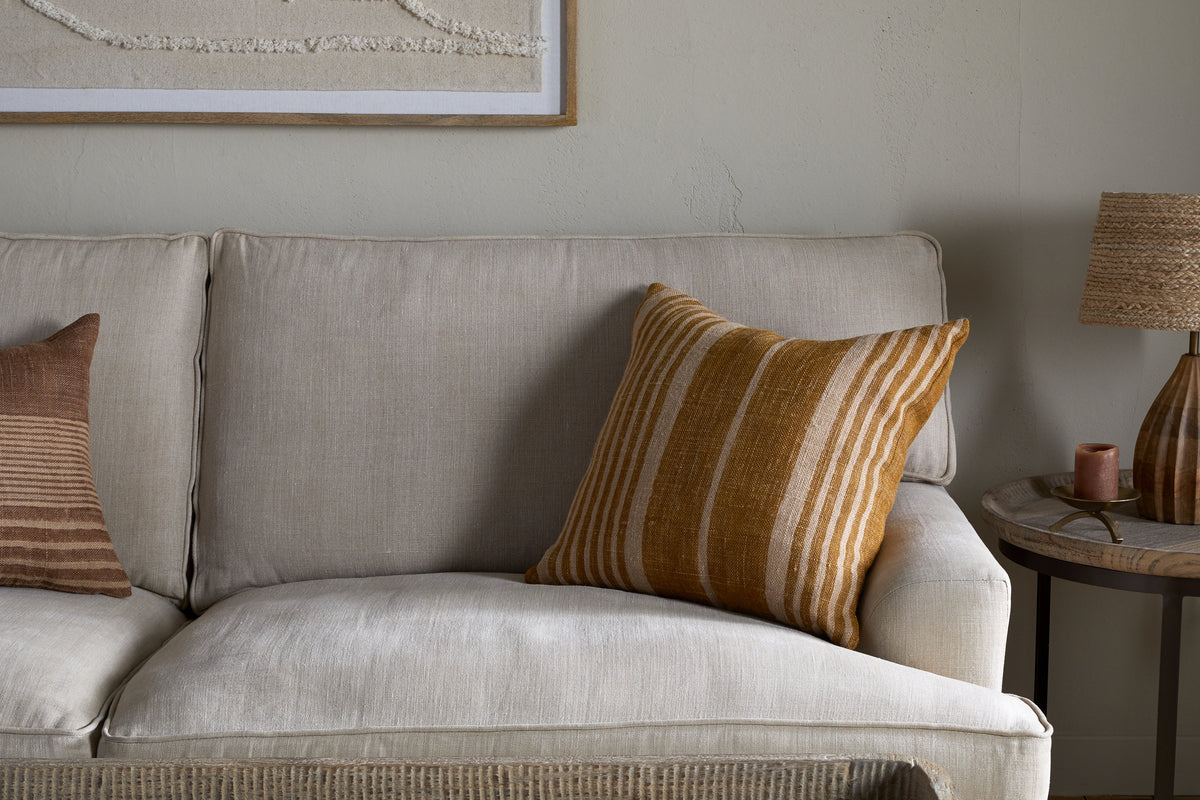 A beige fabric sofa is styled with nkuku’s Eesha Jute Cushion Cover in ochre. Against a light wall, it pairs with a striped brown cushion. A round wooden side table holds a textured lamp and candle beneath minimalist wall art.