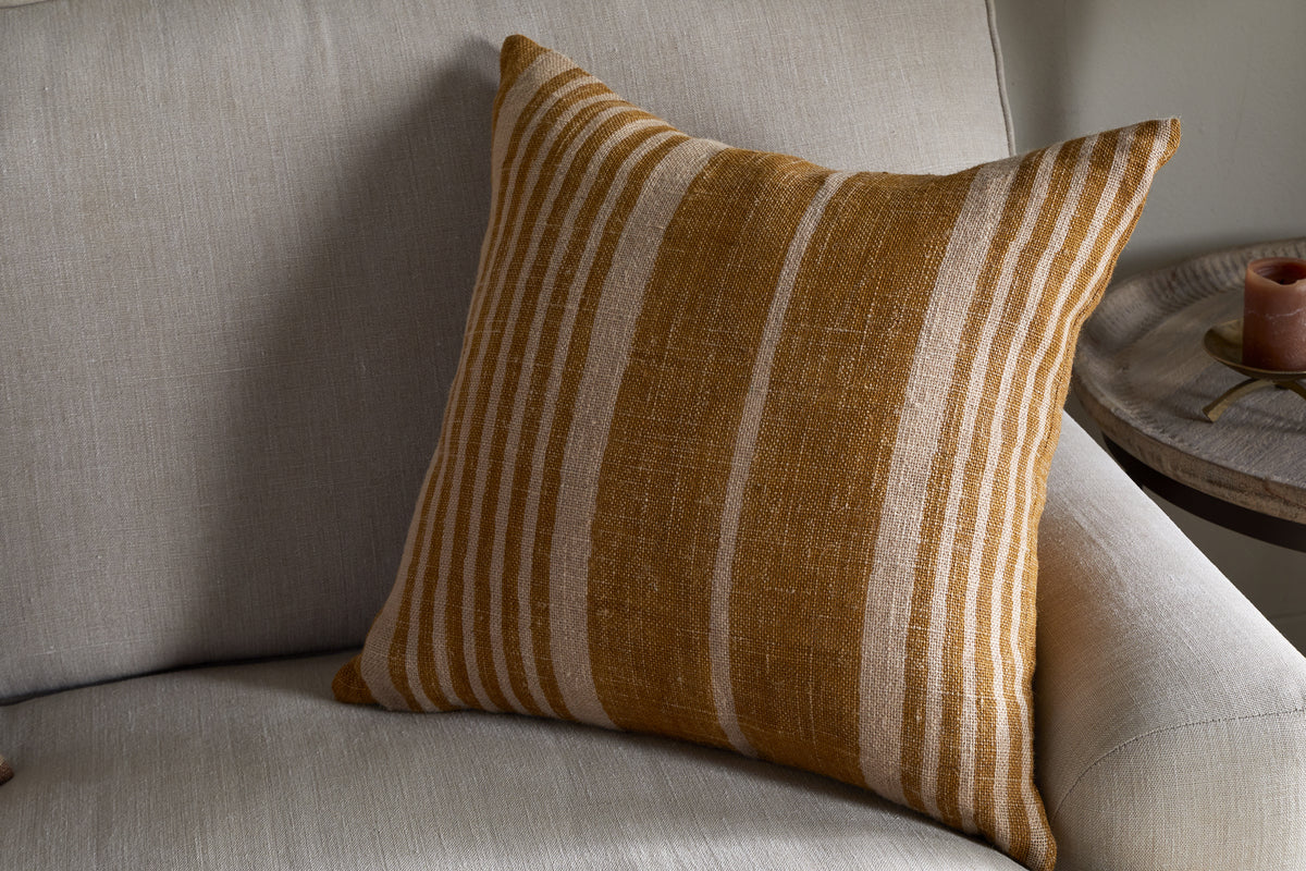 The nkuku Eesha Jute Cushion Cover - Ochre, featuring vertical mustard yellow and beige stripes, adds modern rustic charm to a light gray sofa. A round side table with a brown candle completes the cozy setting.