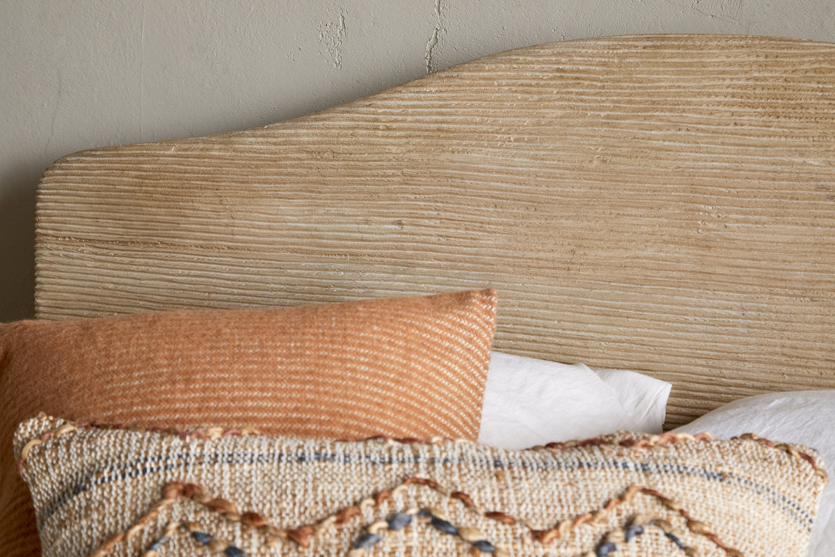 A close-up of the nkuku Vivan Grooved Bed - Natural’s textured wooden headboard behind two pillows—one rust-striped, the other beige with blue and tan embroidery—set against a light wall with visible cracks.