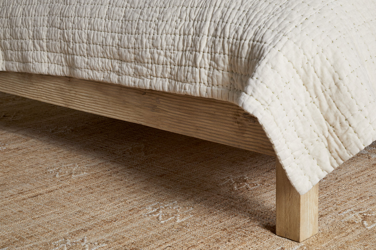 A close-up of the nkuku Vivan Grooved Bed - Natural shows its solid wood frame with visible grain. The bed is topped with a cream quilted blanket and sits on a beige patterned rug, creating a cozy, minimal look.