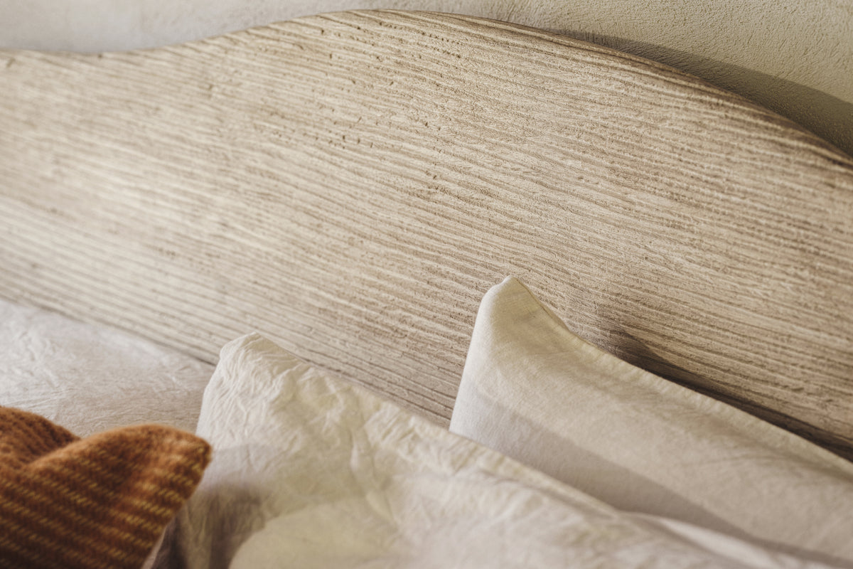 A close-up of the nkuku Vivan Grooved Bed - Natural shows its textured, horizontal-grain headboard. White pillows and sheets with a folded rust knit blanket complete the warm, inviting scene in gentle natural light.