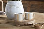 The nkuku Patthar Mug - Cream & Sand - Small (Set of 2) features two striped mugs on a rustic wooden stand, displayed with a white textured vase, woven coasters, and a small bowl of brown sugar on the wooden table.