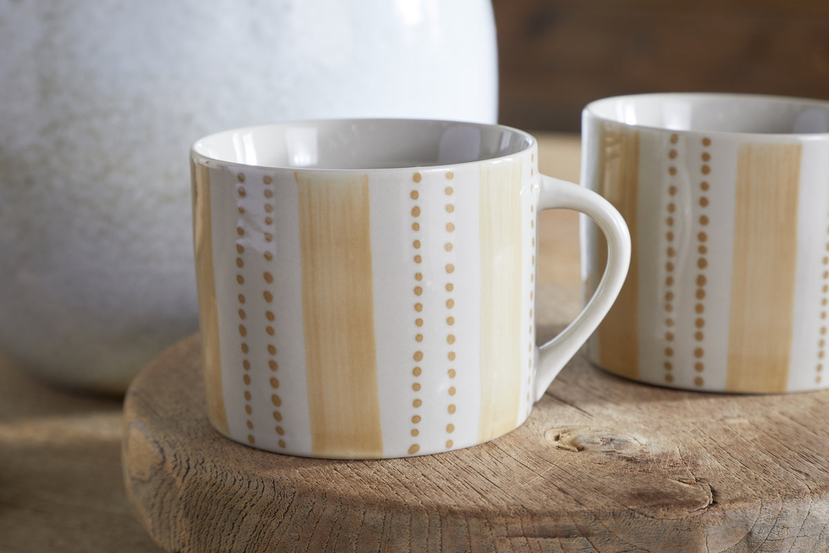The nkuku Patthar Mug - Cream & Sand - Small (Set of 2) features hand-painted stoneware mugs with vertical tan stripes and dotted lines, shown on a round wooden surface in warm, natural light for a cozy, rustic feel.