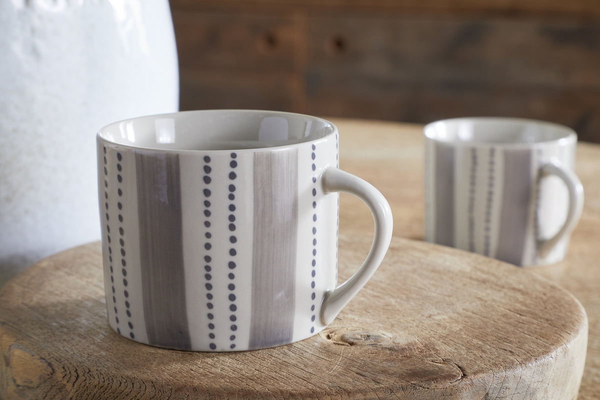 The nkuku Patthar Mug - Cream & Grey - Small (Set of 2) features hand-painted vertical gray stripes and dotted lines, shown on a wooden surface with another in the background, evoking a cozy, rustic vibe with muted tones and soft lighting.