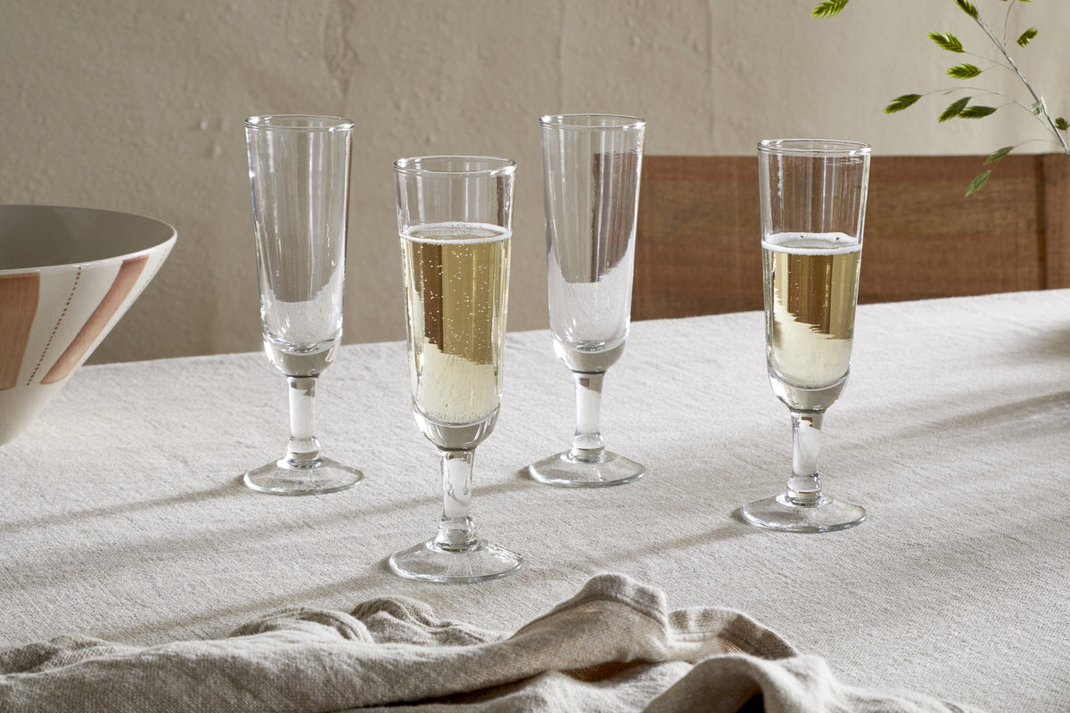 Four Pani Champagne Flutes by nkuku, in a set of 4 clear glasses, are arranged on a beige tablecloth; two are filled with bubbly champagne and two are empty. A folded napkin rests nearby alongside a striped bowl and leafy branch.