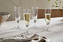Four Pani Champagne Flutes by nkuku, in a set of 4 clear glasses, are arranged on a beige tablecloth; two are filled with bubbly champagne and two are empty. A folded napkin rests nearby alongside a striped bowl and leafy branch.