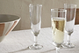 Four nkuku Pani Champagne Flute glasses in clear recycled glass are displayed on a beige tablecloth, three empty and one filled with bubbly, next to a striped ceramic bowl against a plain cream wall.