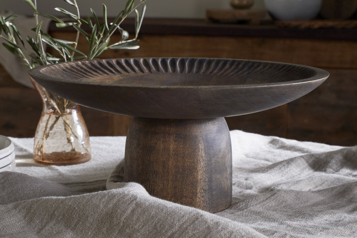 The nkuku Eisha Wooden Cake Stand in brown, featuring a hand-carved wide, shallow top with a fluted rim, sits on a textured cloth. In the background, a small glass vase with green leaves adds rustic charm to the scene.
