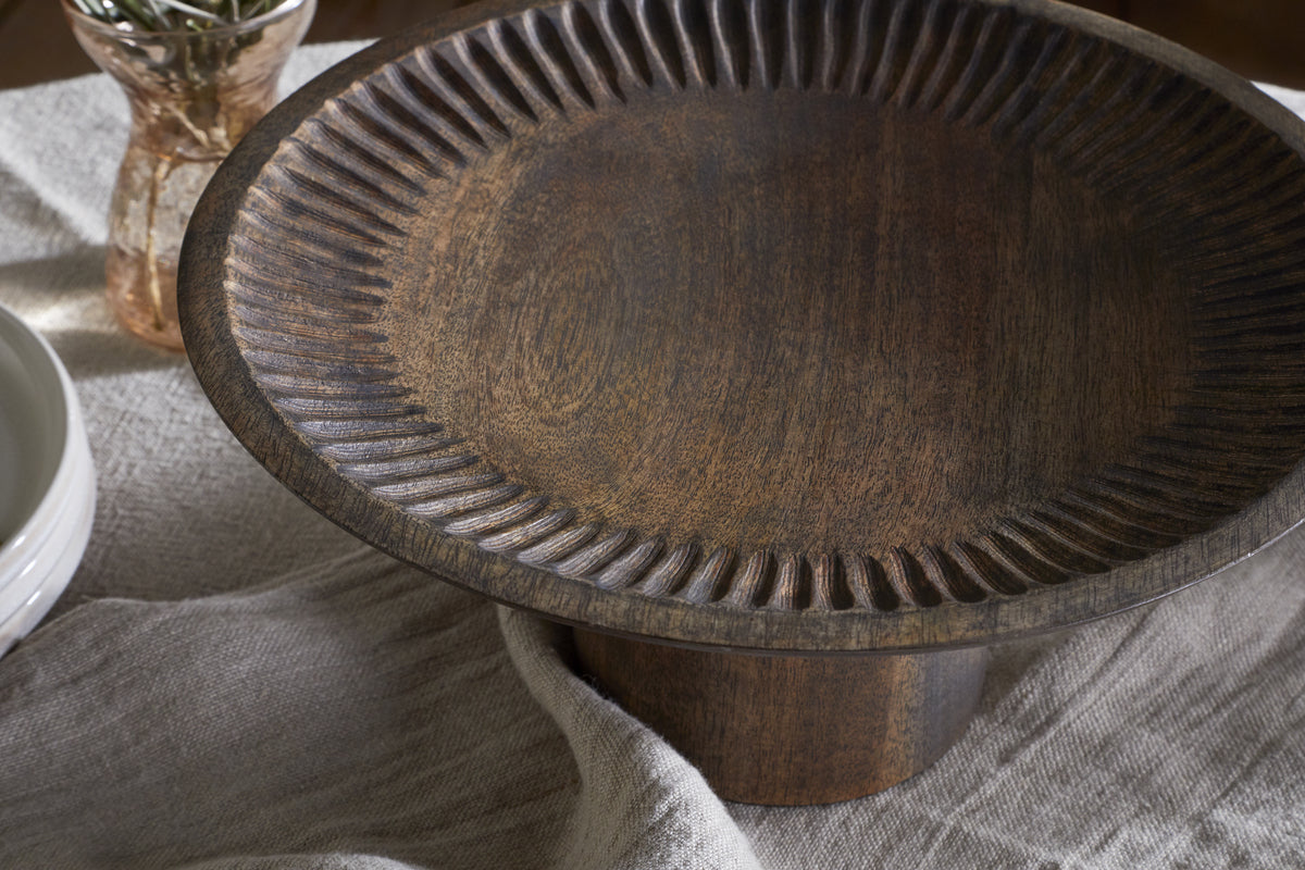 The Eisha Wooden Cake Stand - Brown by nkuku, a hand-carved dark wood pedestal, sits atop textured fabric. Nearby, a small vase and stacked white dishes enhance the rustic, natural aesthetic—ideal for cakes or serveware.