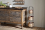 A rustic kitchen showcases a wooden cabinet, jars, a plant, and wall shelf. Next to it stands the nkuku Montali Iron & Rattan Three-Tiered Storage Stand in Natural, neatly holding baskets of assorted fruits and vegetables.