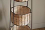 The nkuku Montali Iron & Rattan Three-Tiered Storage Stand - Natural features round handwoven baskets on a dark metal frame, ideal for kitchen or pantry organization. Shown on a wooden floor against a textured beige wall.