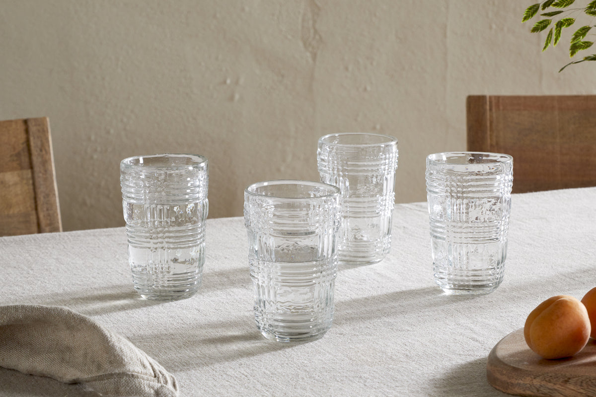 The Kaanch Highball (Set of 4) by nkuku, featuring clear textured glass, is arranged on a light tablecloth with wooden chairs, a wooden plate with apricots, green leaves, and a folded napkin for a fresh look.
