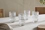 A set of four nkuku Kaanch Highball glasses in clear glass, featuring horizontal ridges, are arranged on a beige tablecloth with wooden chairs and a cutting board softly visible in the background.