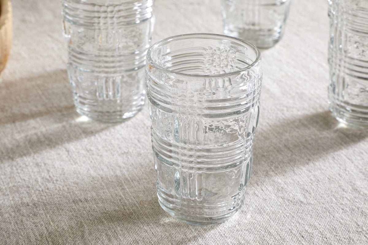 The nkuku Kaanch Highball - Clear (Set of 4) features embossed geometric glass tumblers, displayed on a light beige fabric with one centered in focus and three softly blurred behind.
