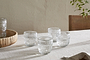 Four nkuku Kaanch Tumblers - Clear (Set of 4) with an embossed check pattern rest on a light tablecloth. A wooden tray with another Kaanch tumbler is on the left, and a chair with green stems appears to the right against a smooth wall.